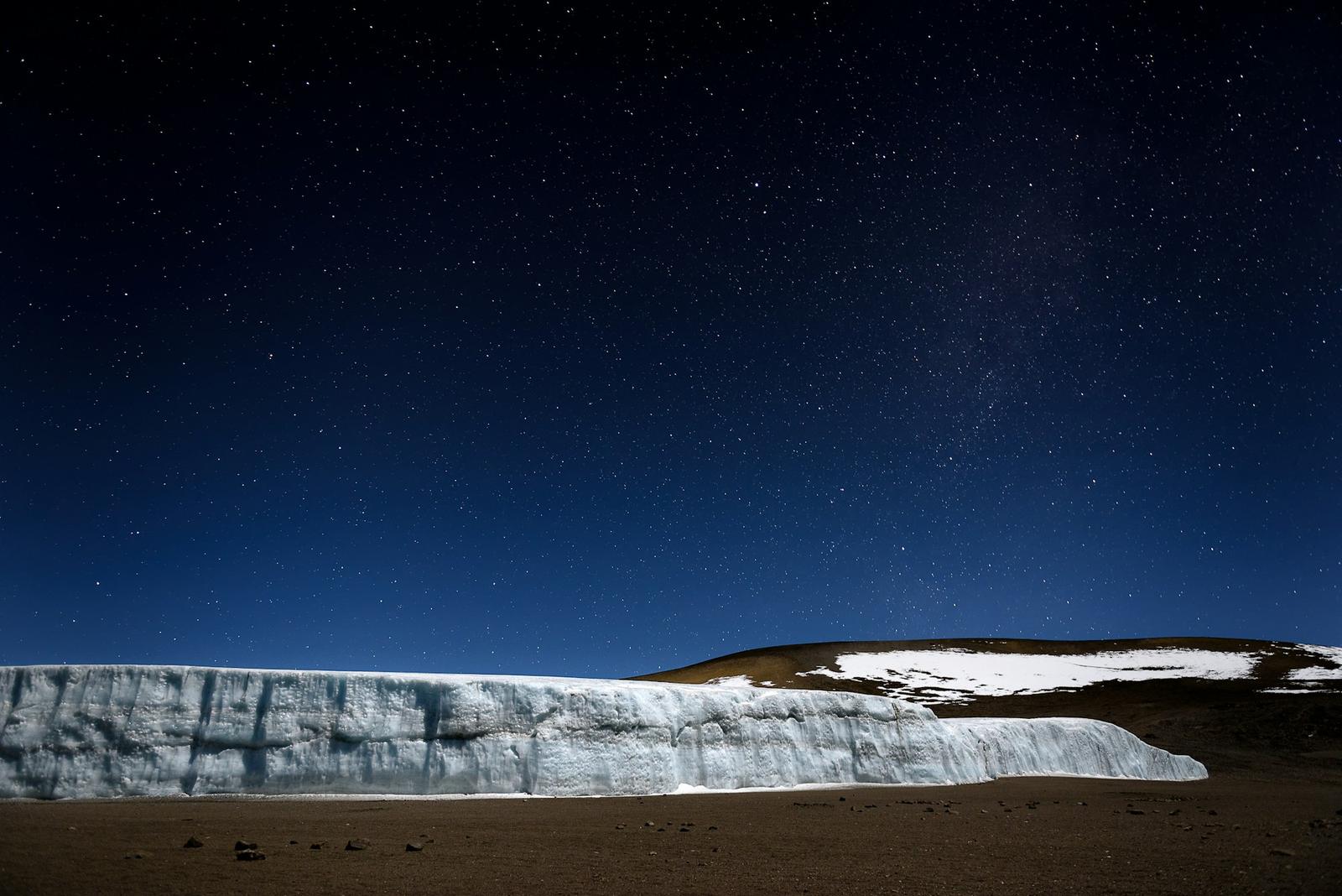 A wall of glacier ice in Kibo’s summit crater under a sky full of stars, photographed at night from Crater Camp